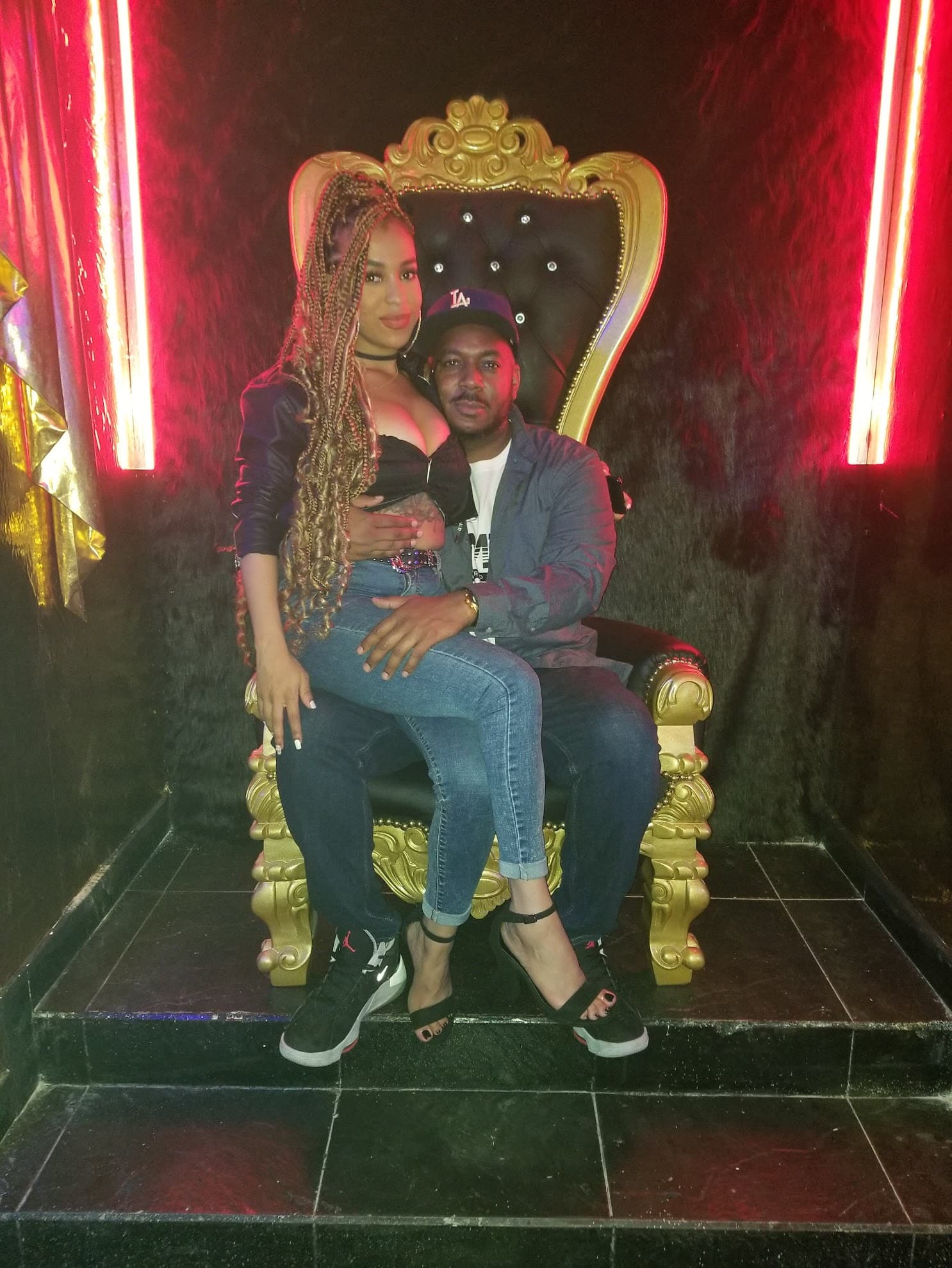 Man and woman posing on an ornate gold throne between two vertical red neon lights.