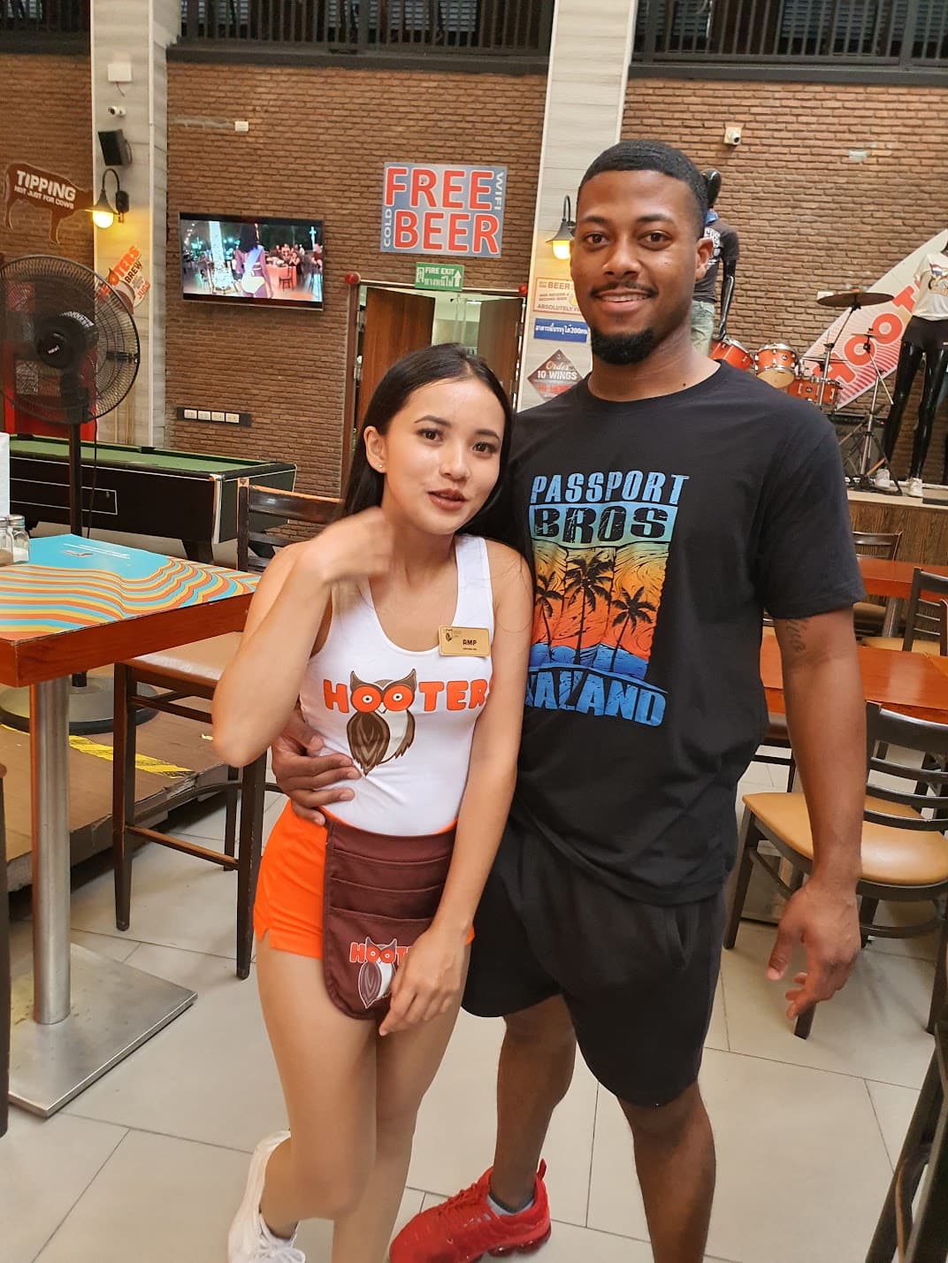 Man in a Passport Bros Thailand shirt posing with a Hooters waitress in a restaurant.