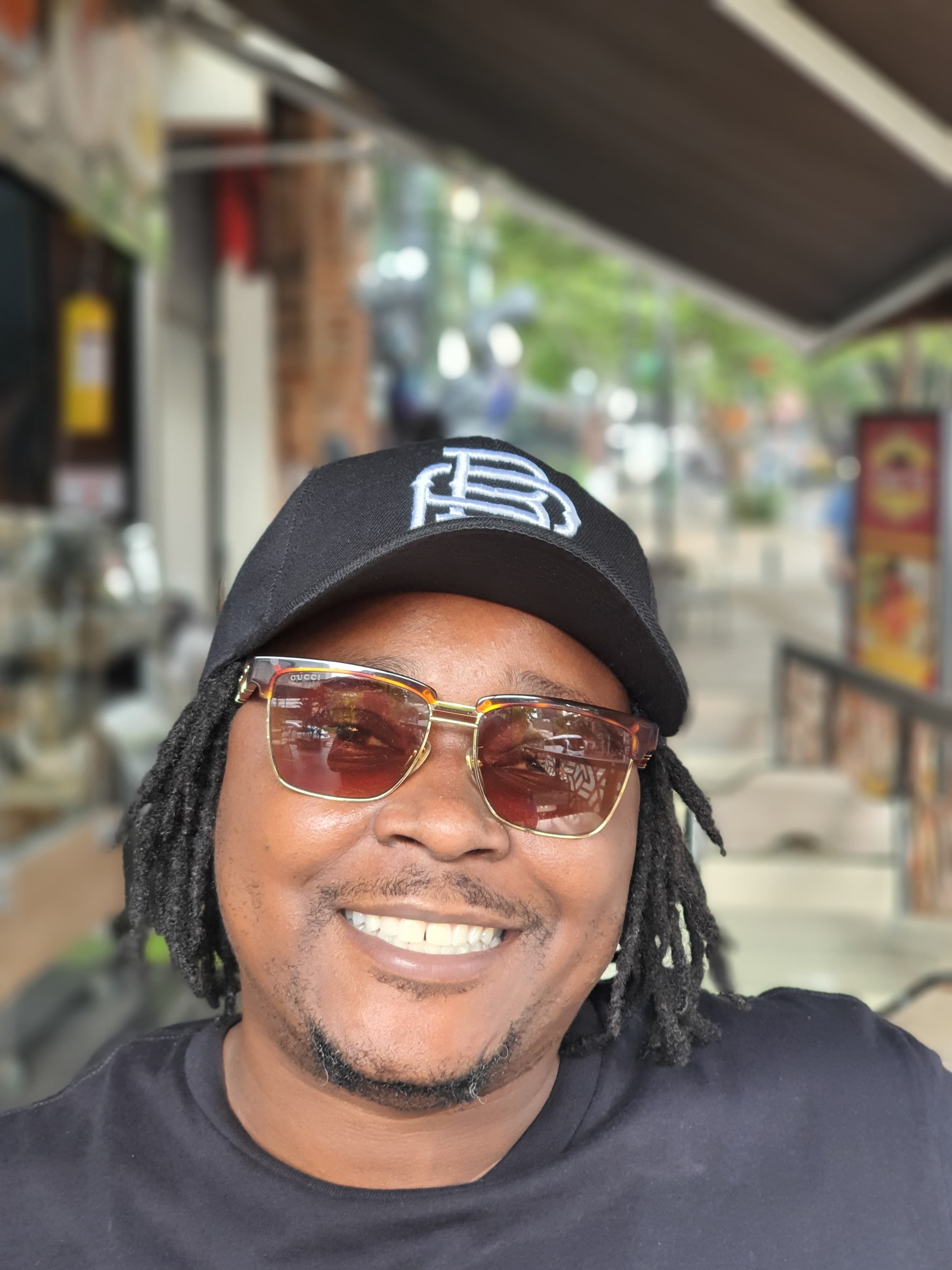 Smiling Black man with dreadlocks wearing a black cap and gold-rimmed sunglasses outdoors.