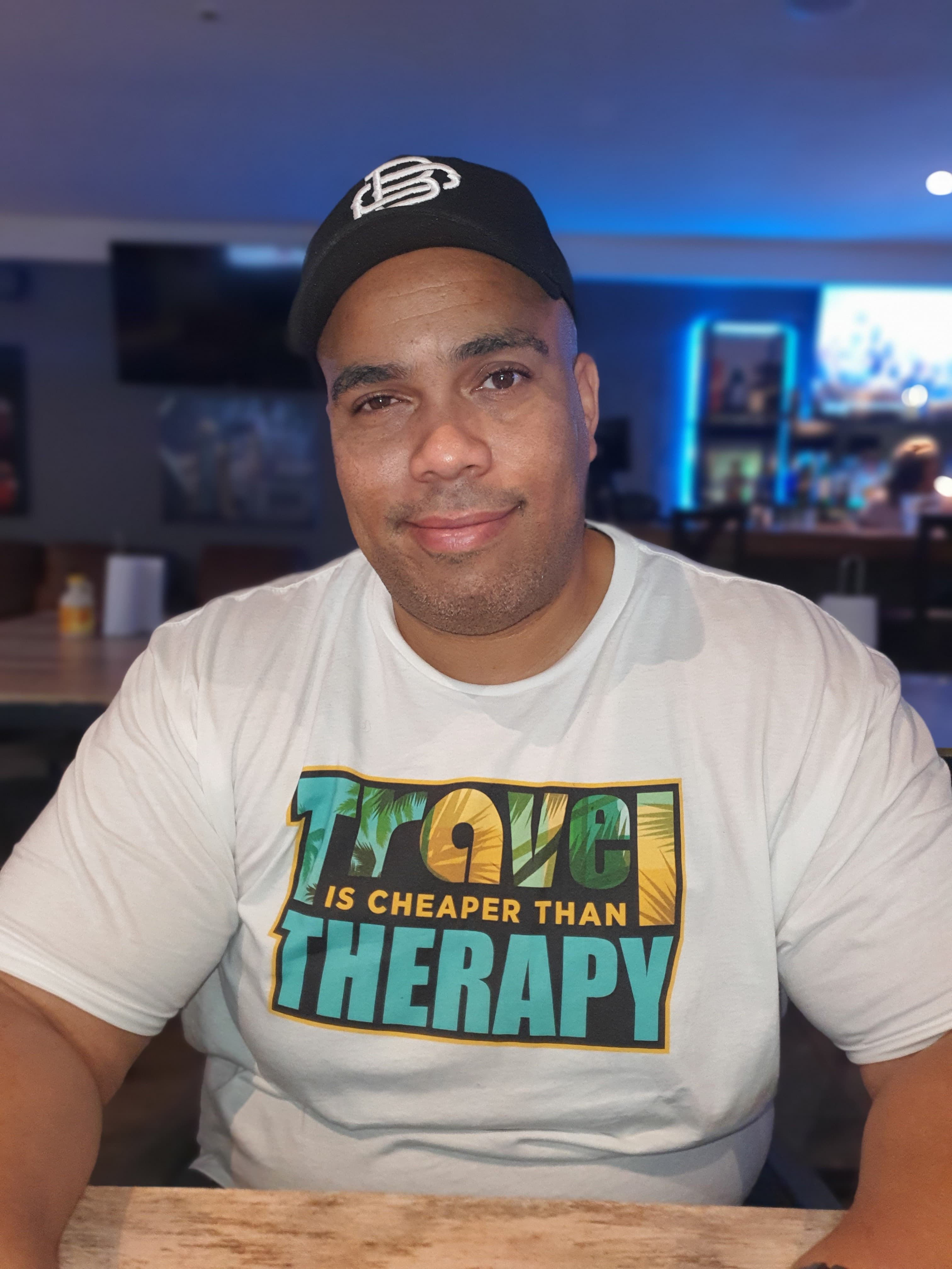 Smiling man in a black cap and white t-shirt reading "Travel is cheaper than therapy."