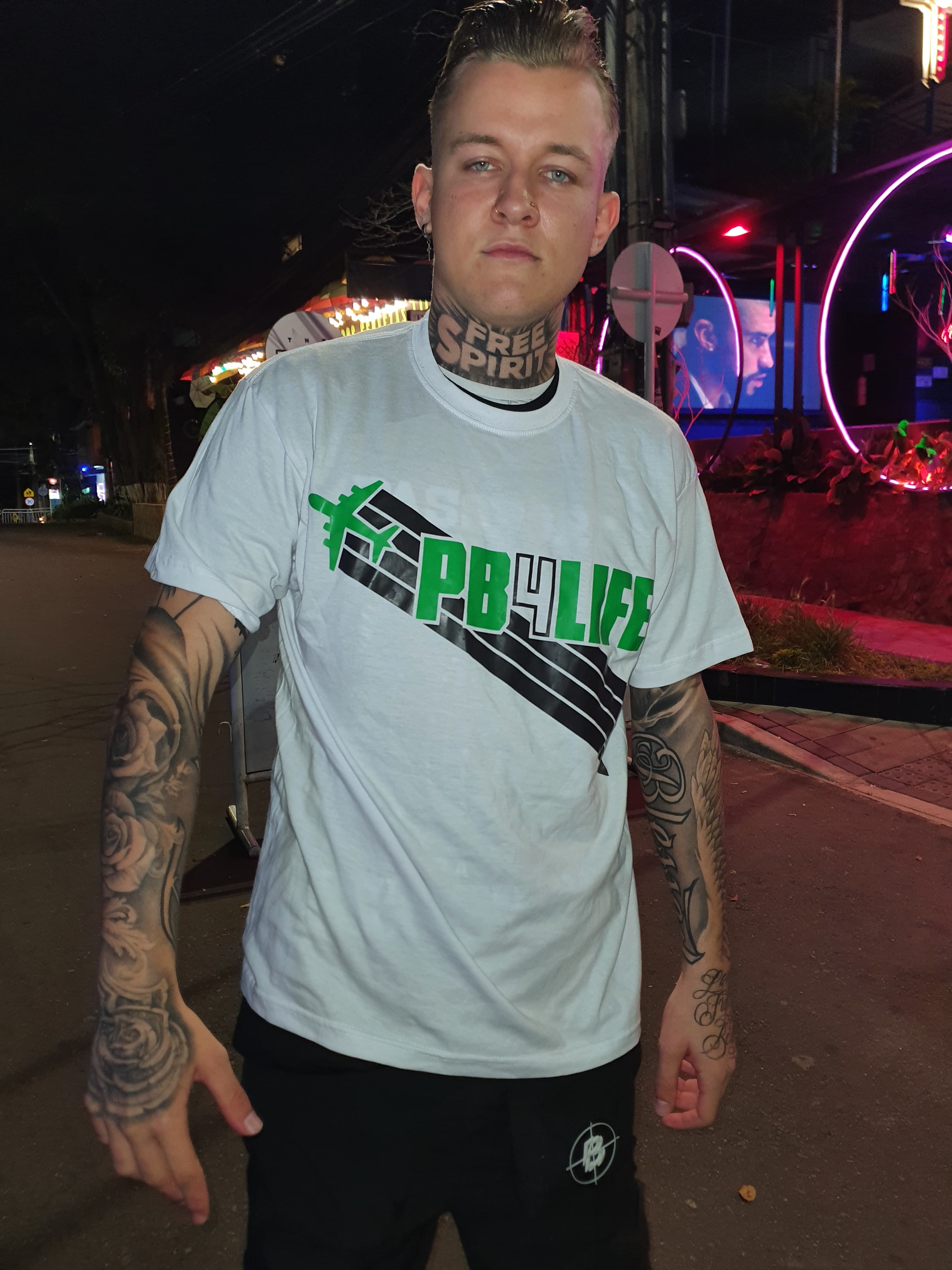 Man with neck and arm tattoos wearing a white PB4LIFE t-shirt under neon lights.