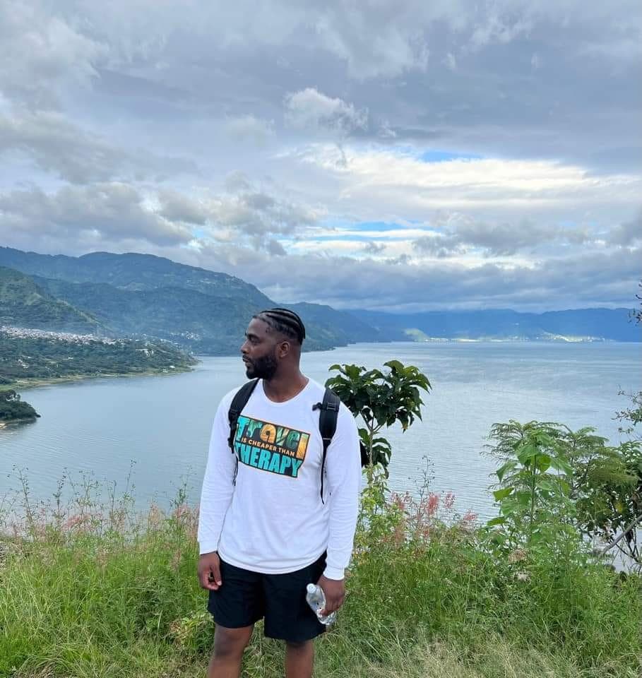 Bearded man in a travel shirt overlooks a scenic lake and mountains under cloudy skies.