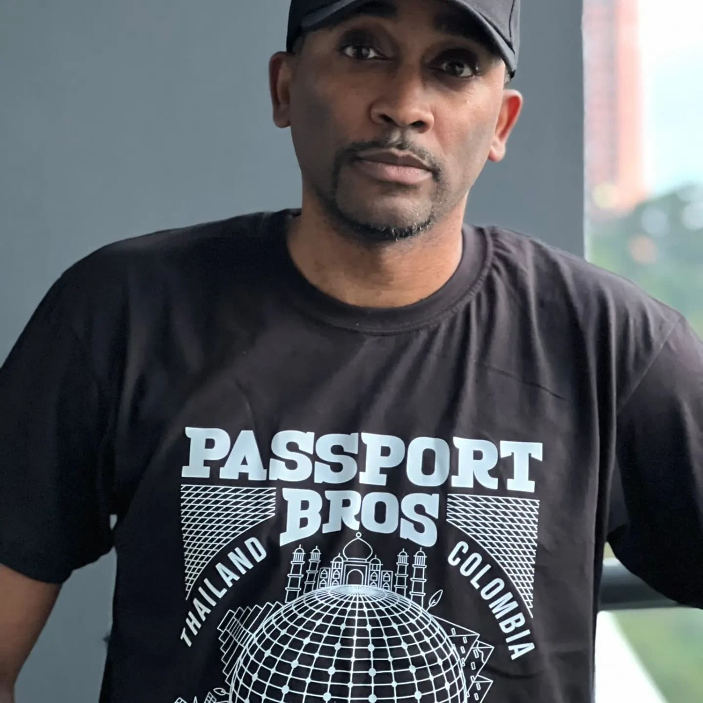 Man wearing a black cap and Passport Bros shirt with Thailand and Colombia graphics.