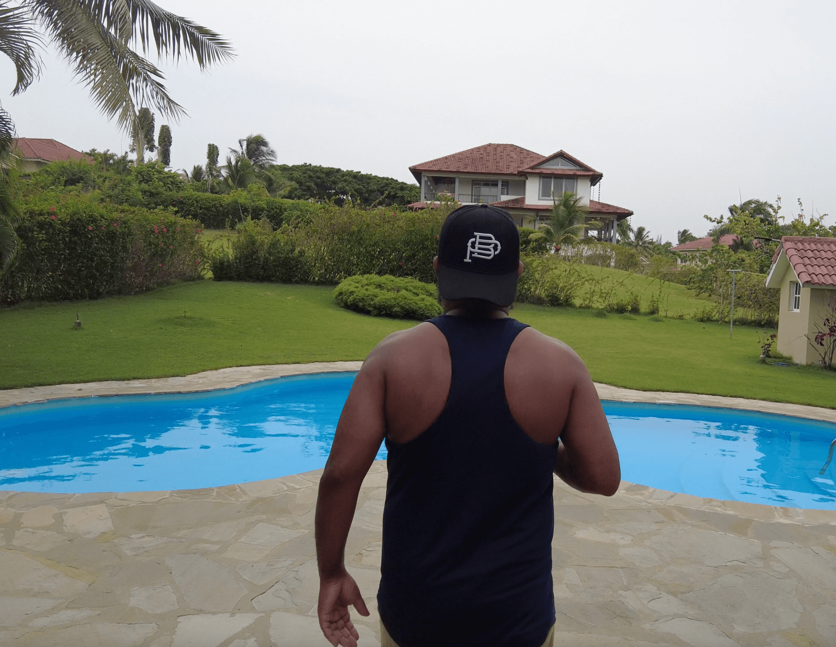 Man in black tank top and cap stands by a blue pool with tropical house.