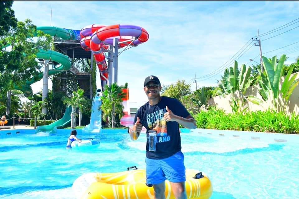 Smiling man giving thumbs up in a water park pool with colorful slides in the background.