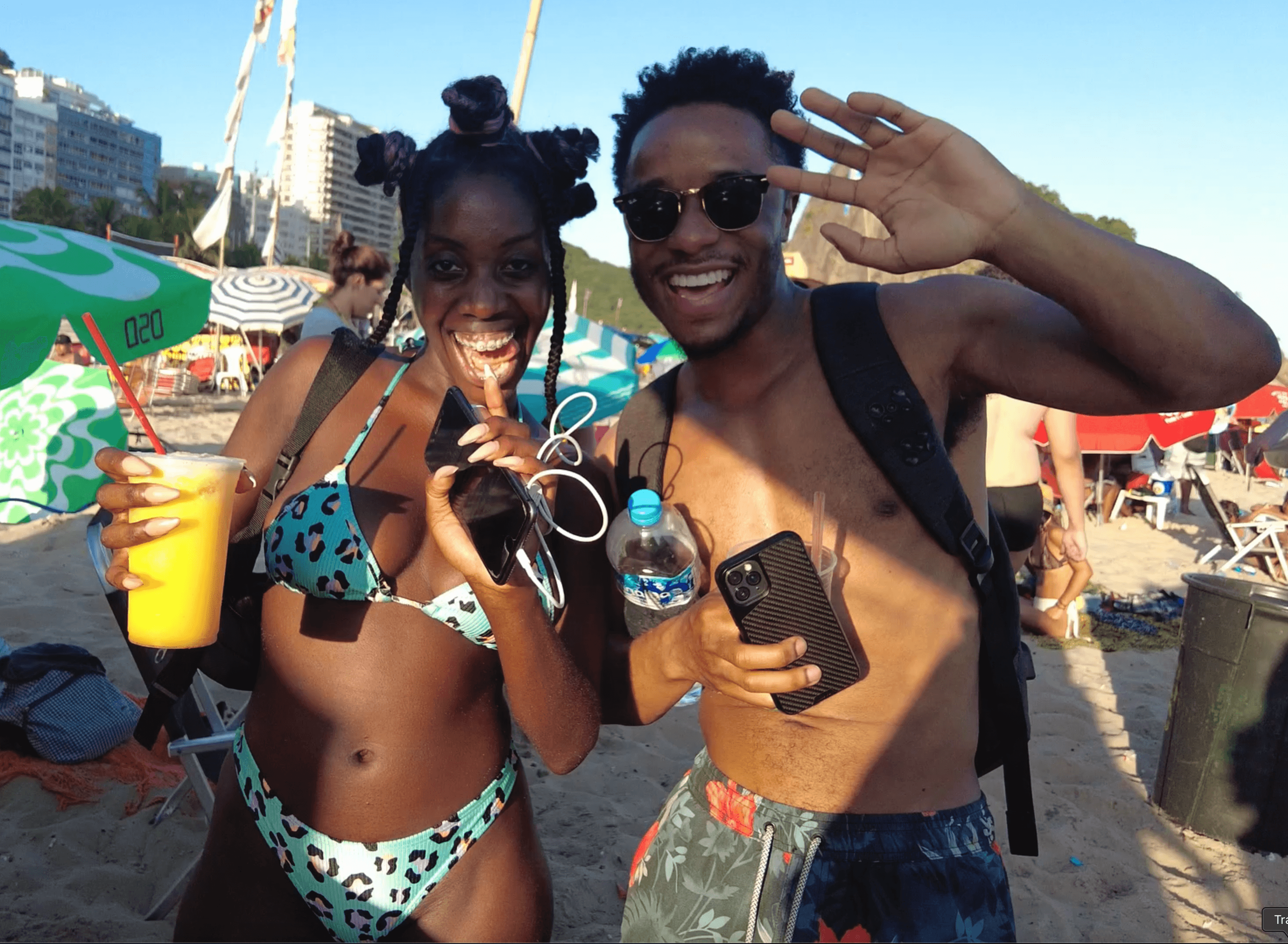 Two smiling Black people in swimwear posing on a sunny beach, holding drinks and waving.