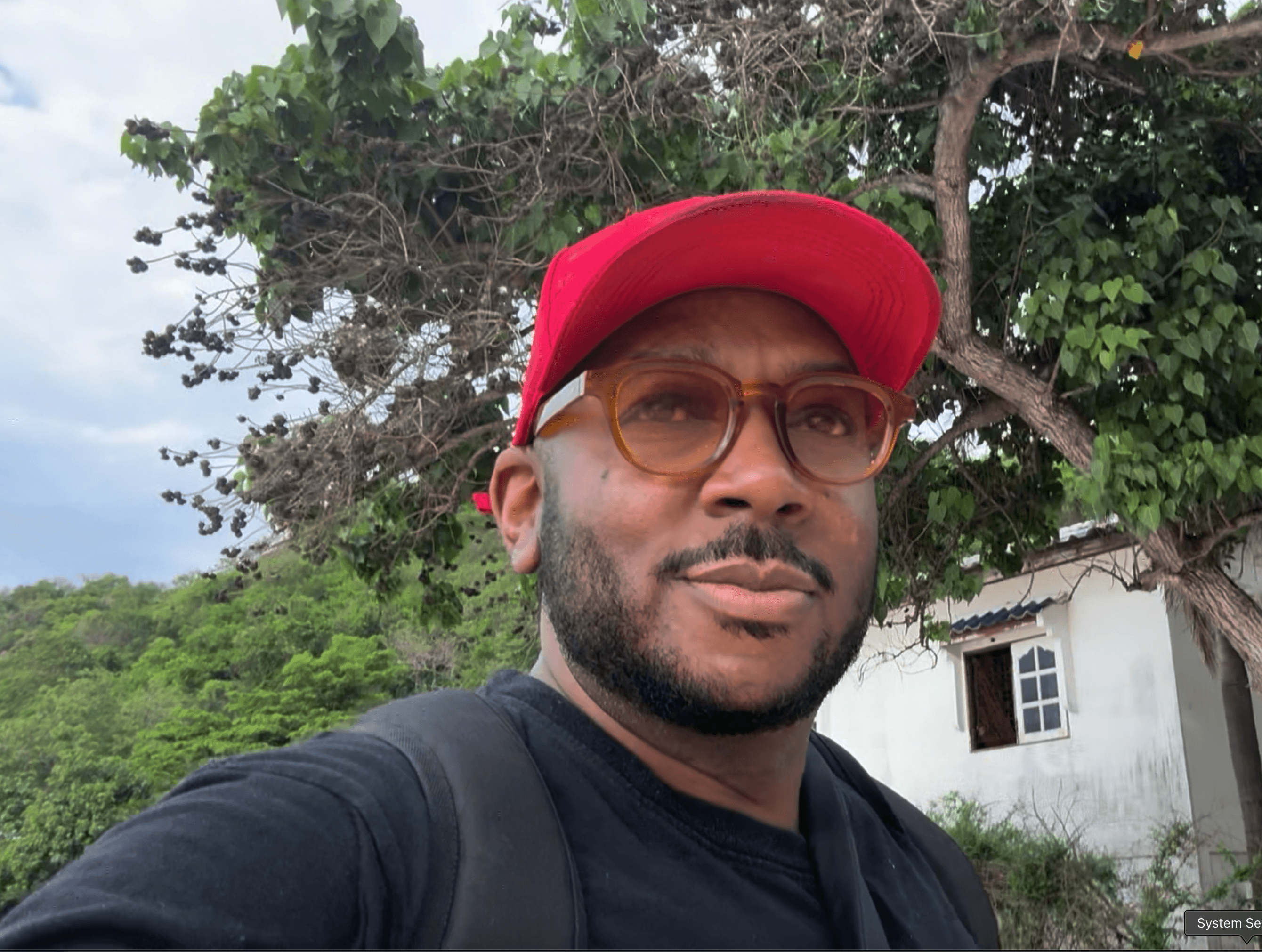 Bearded man wearing a red cap and amber glasses outdoors near a white building.