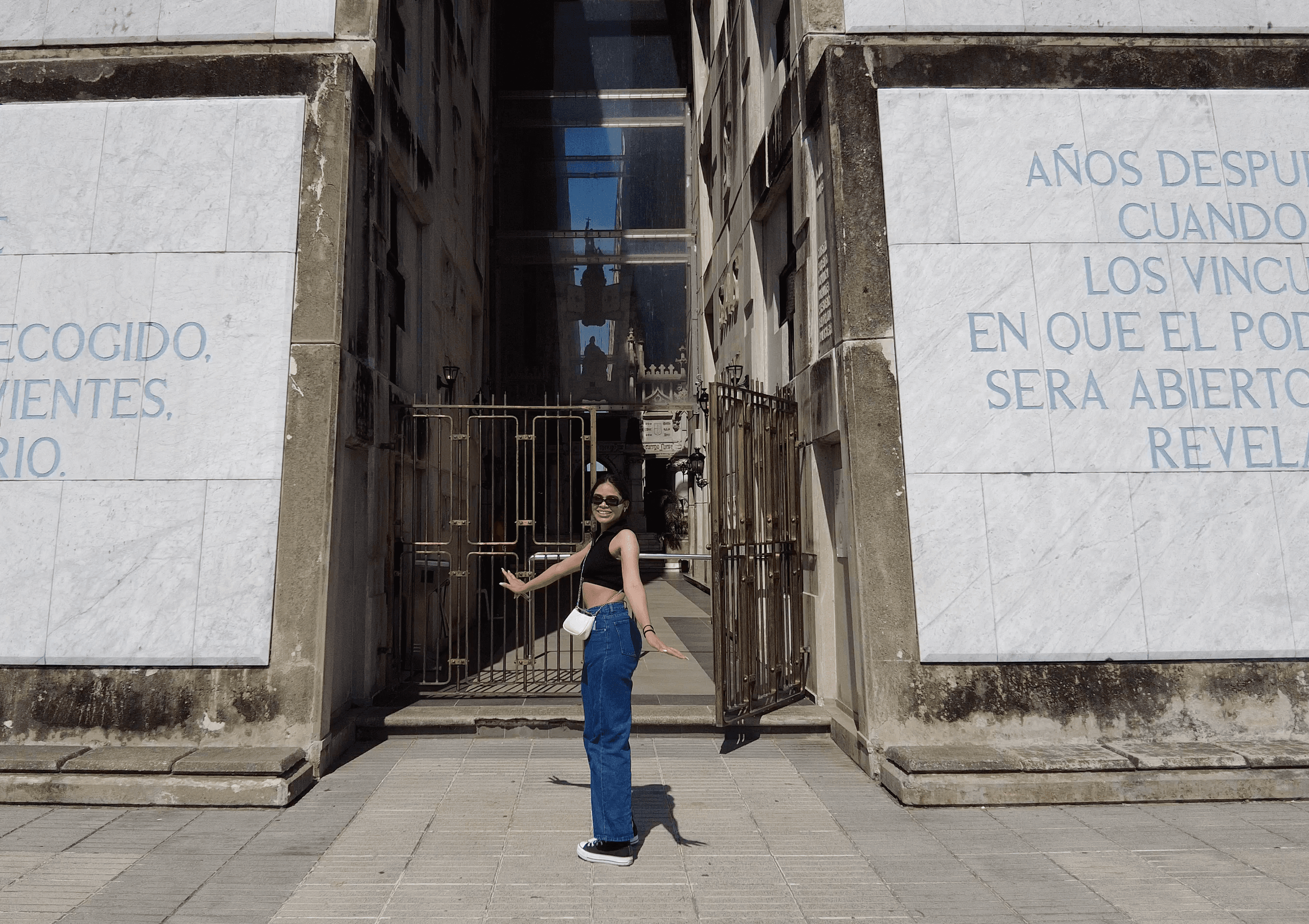 Smiling woman in jeans poses before a stone monument with Spanish inscriptions and metal gates.