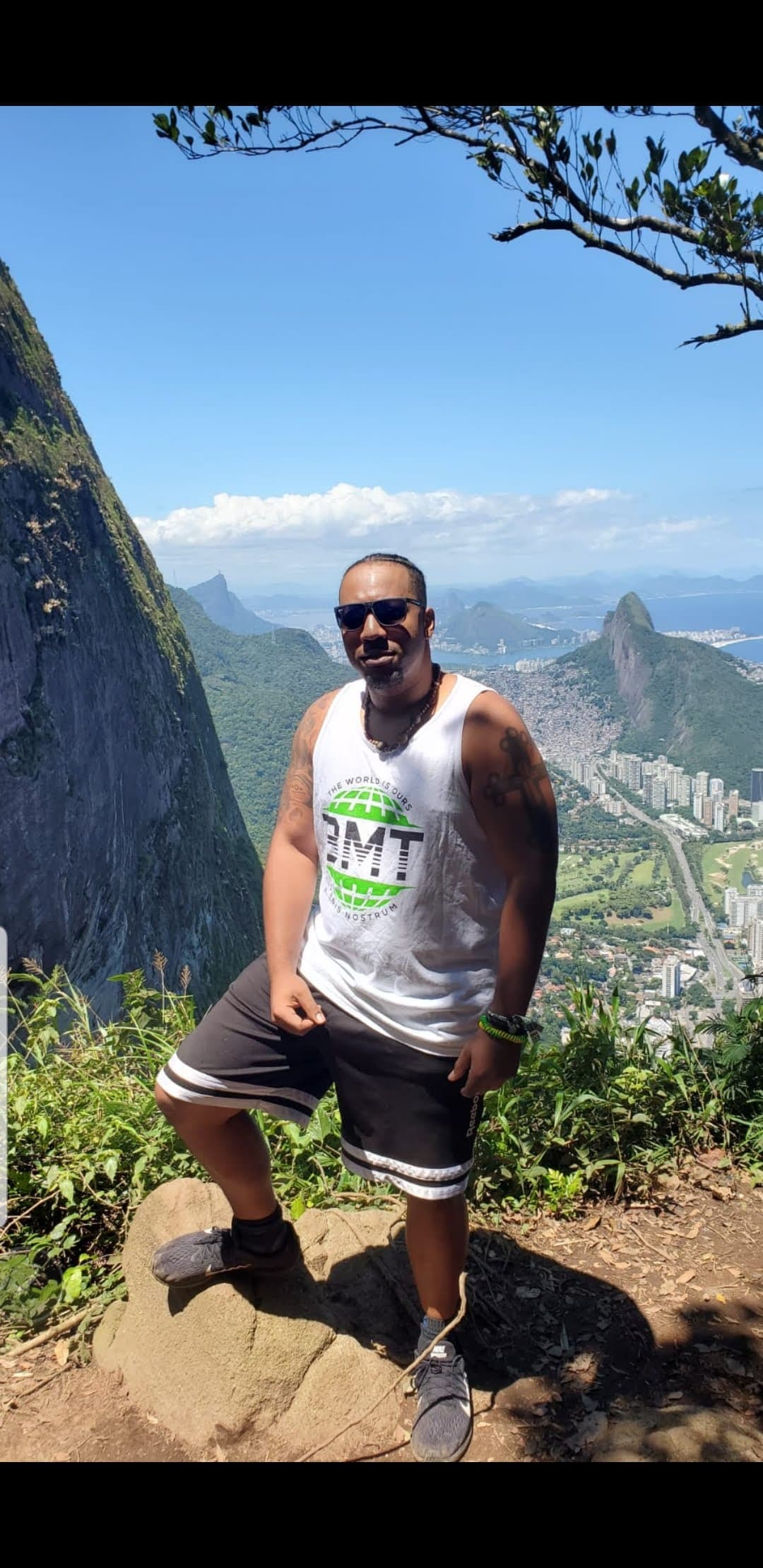 Man in a white tank top poses on a rocky peak overlooking a coastal city.