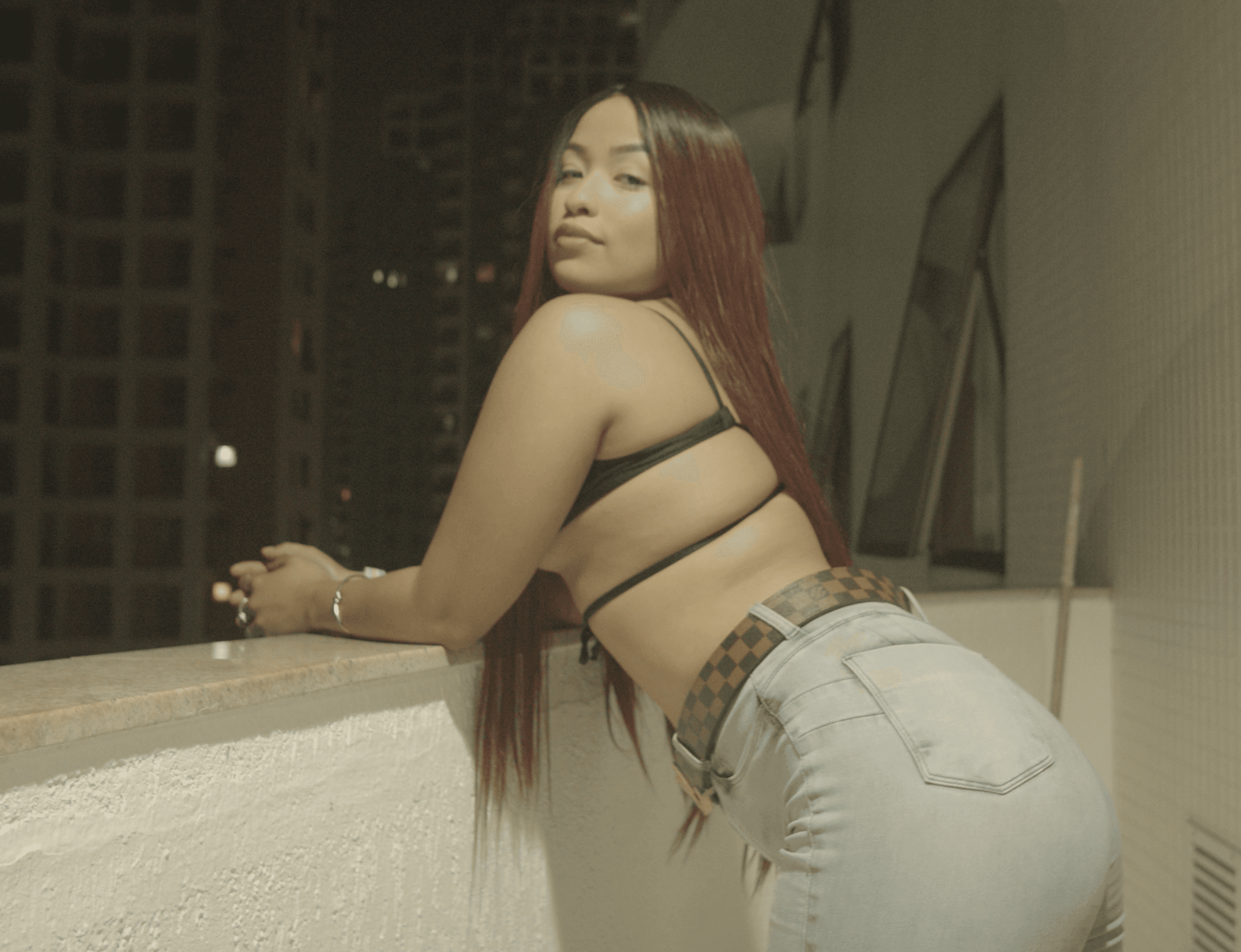 Red-haired woman in jeans and strappy top leans on a balcony railing at night.
