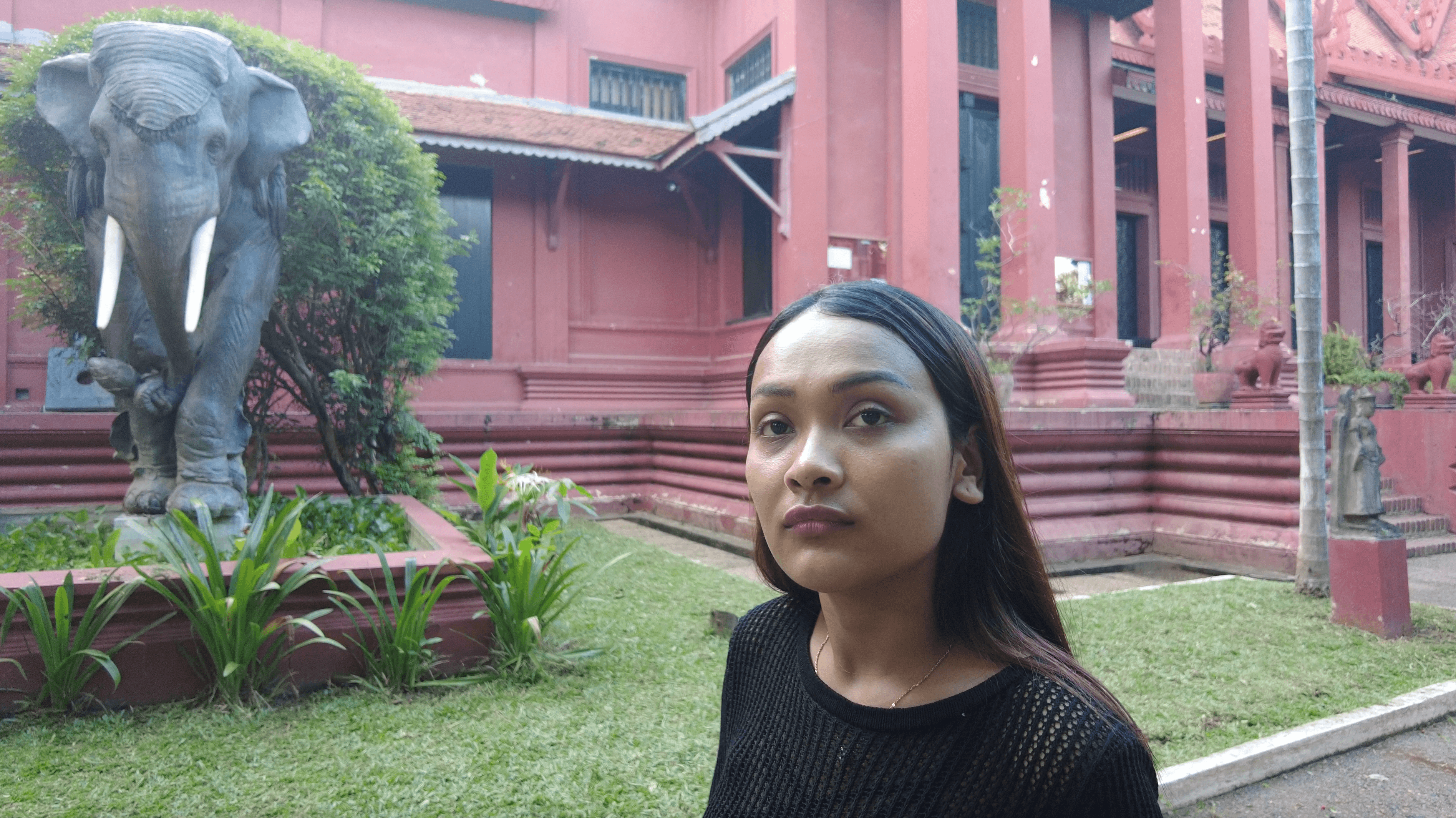A woman stands before a large elephant statue and a red traditional museum building.