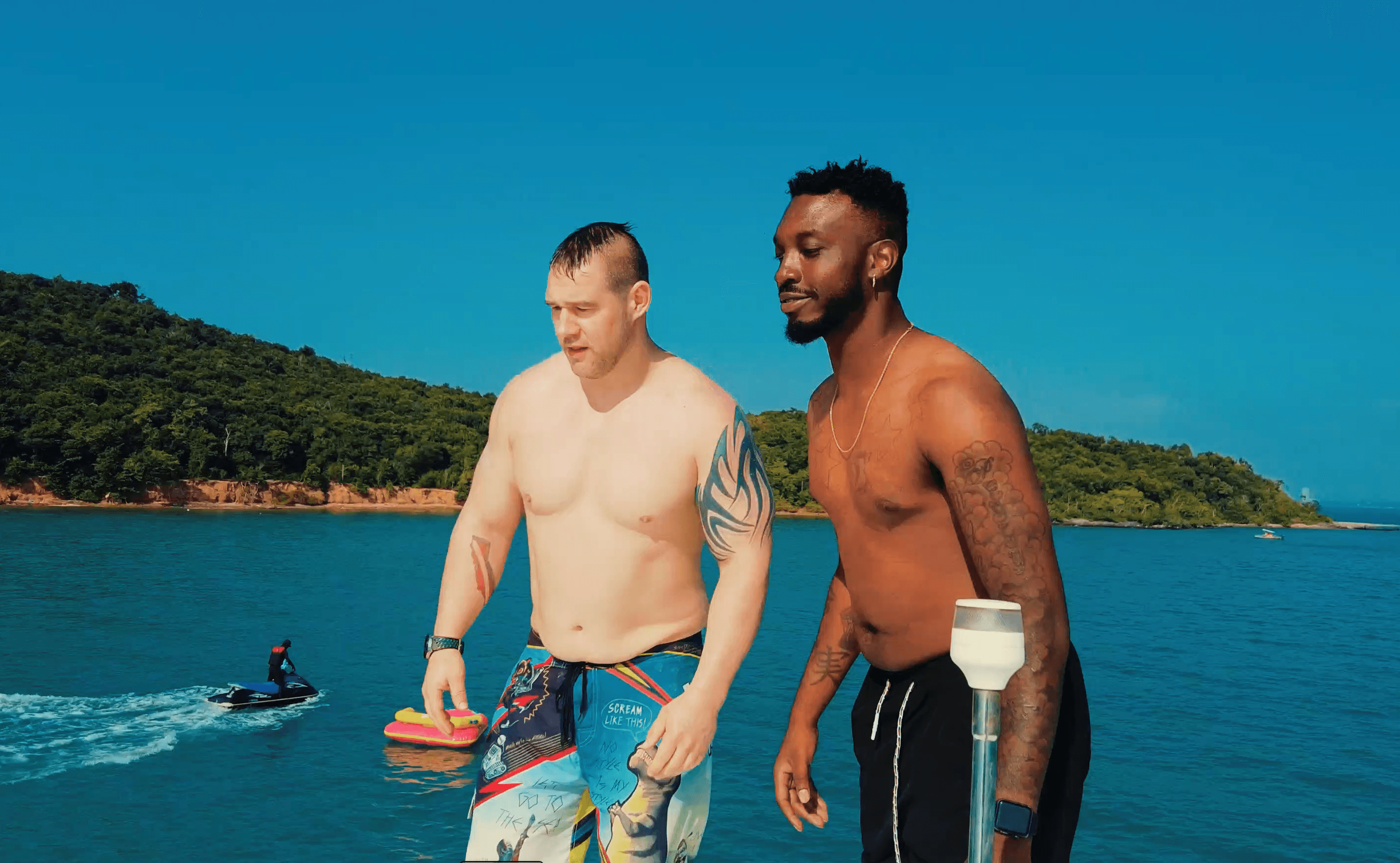 Two shirtless tattooed men stand on a boat overlooking blue water and a lush coastline.